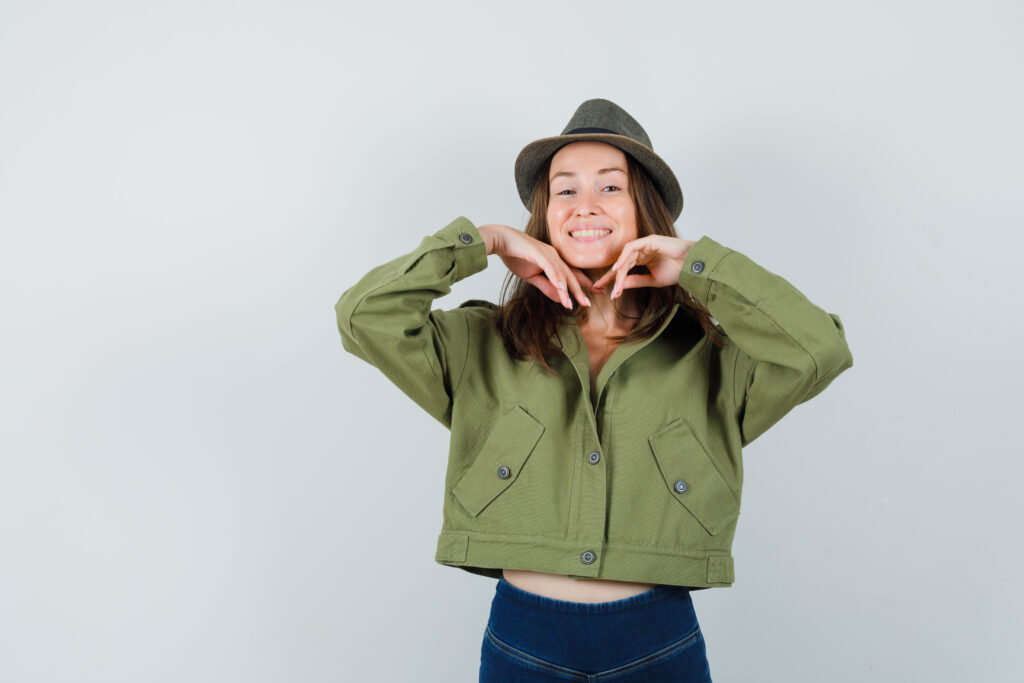 Young female keeping hands touching chin in jacket, pants, hat and looking graceful , front view.